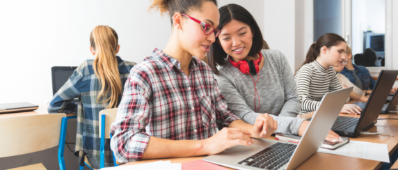 Two young adults in a classroom looking at the screen of a laptop cumputer and smiling