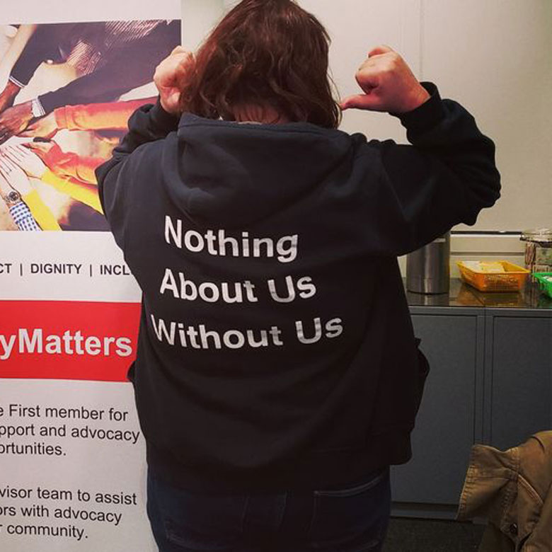 Person standing backwards and pointing to the back of their hoodie, which says